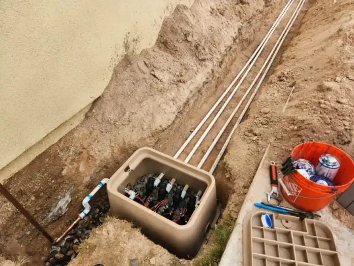 Open irrigation valve box with multiple valves, tools, and white pipes along a trench